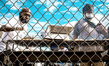 two men cooking on a grill through a chain link fence