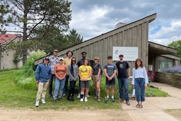 Students standing outside in front of an A-frame building during the summer. 